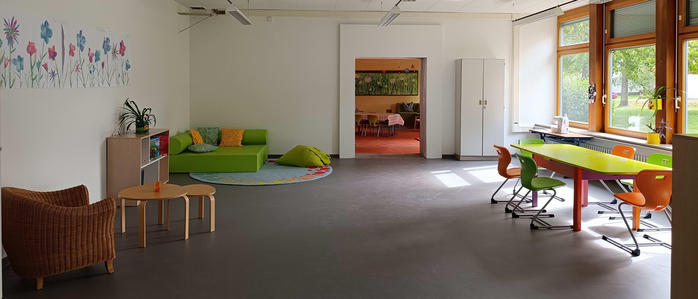 Gemütlicher Aufenthaltsraum mit Korbsessel, kleinen Holztischen, Bücherregal und grünen Sitzmöbeln unter einem bunten Blumenbild