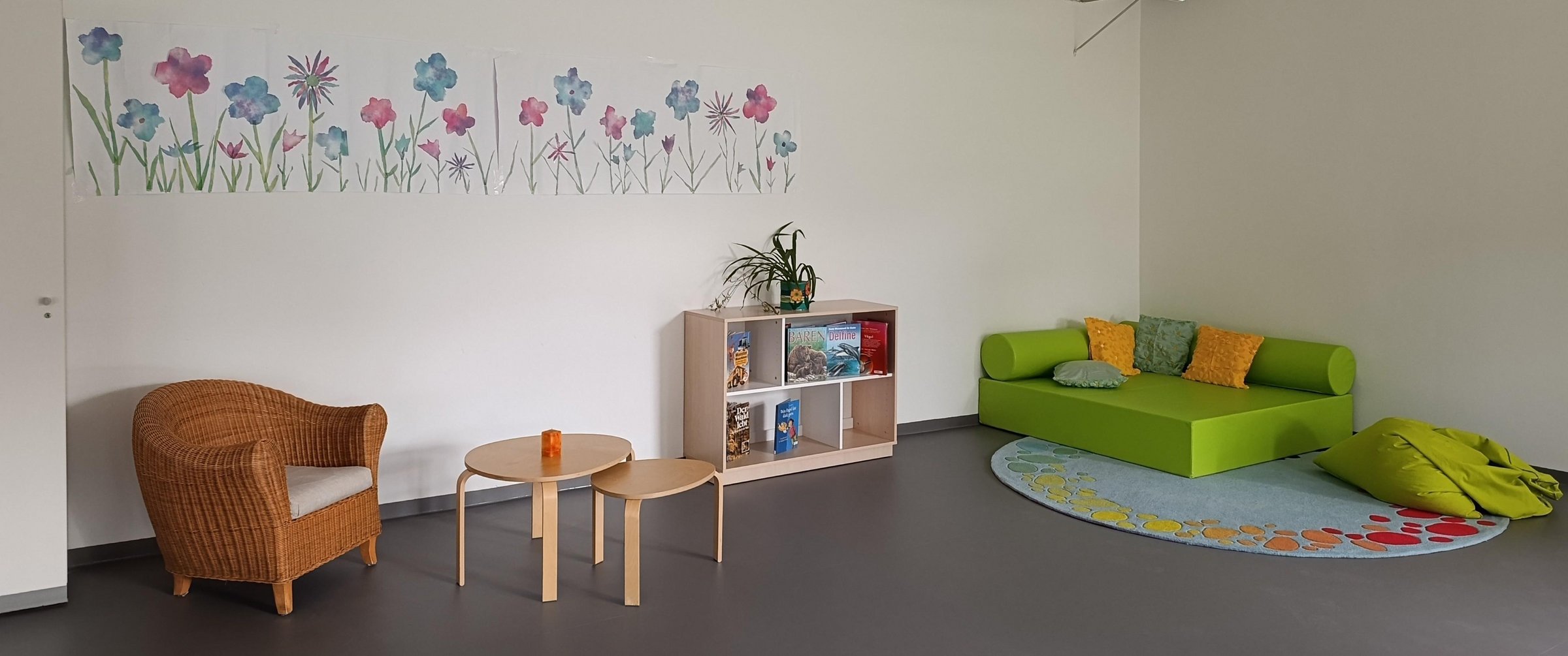 Gemütlicher Aufenthaltsraum mit Korbsessel, kleinen Holztischen, Bücherregal und grünen Sitzmöbeln unter einem bunten Blumenbild